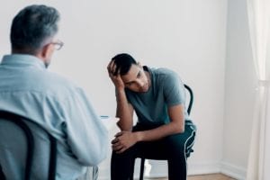 young male patient sits with his head in his hand while a male therapist takes notes in a drug and alcohol detox center