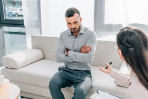 man with crossed arms sits on couch and talks to female therapist in an outpatient rehab center