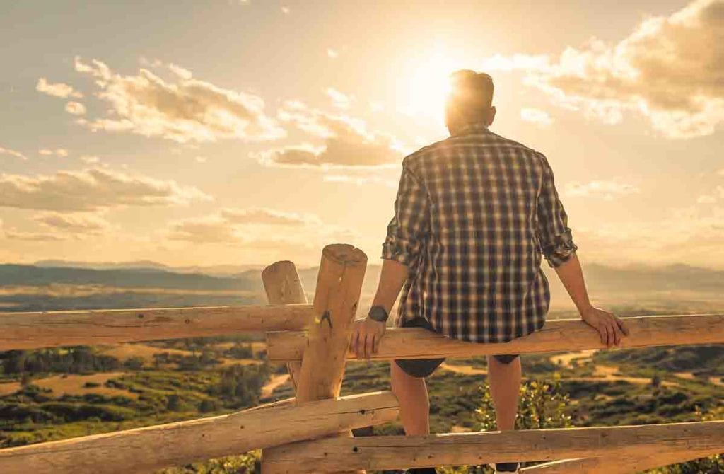 Man sitting on a wooden fence looking towards a sunny landscape at the end of the stages of addiction recovery