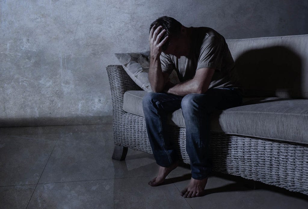 distressed man sitting on couch with head in hands thinking about the stages of alcoholism